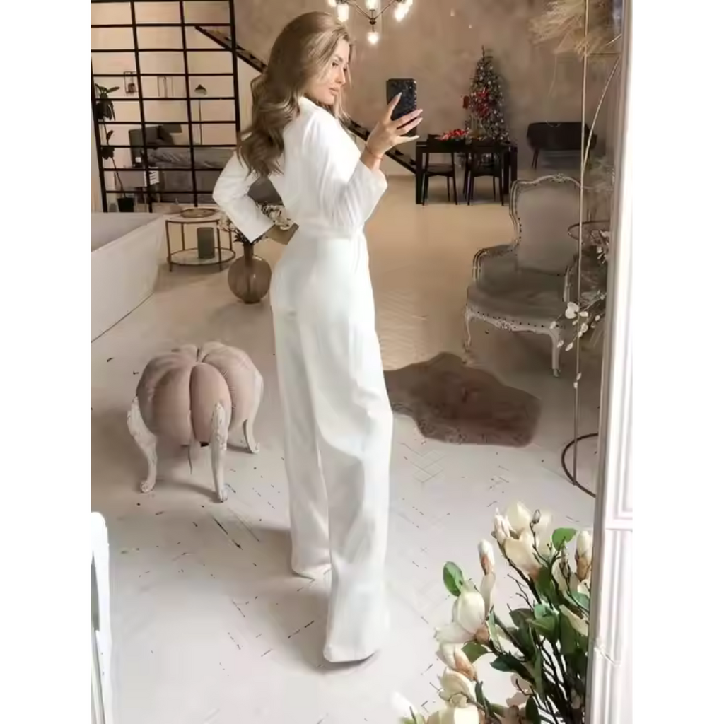 Femme en tenue blanche élégante posant devant un miroir dans un salon chic, décoré avec des meubles vintage et des fleurs blanches.