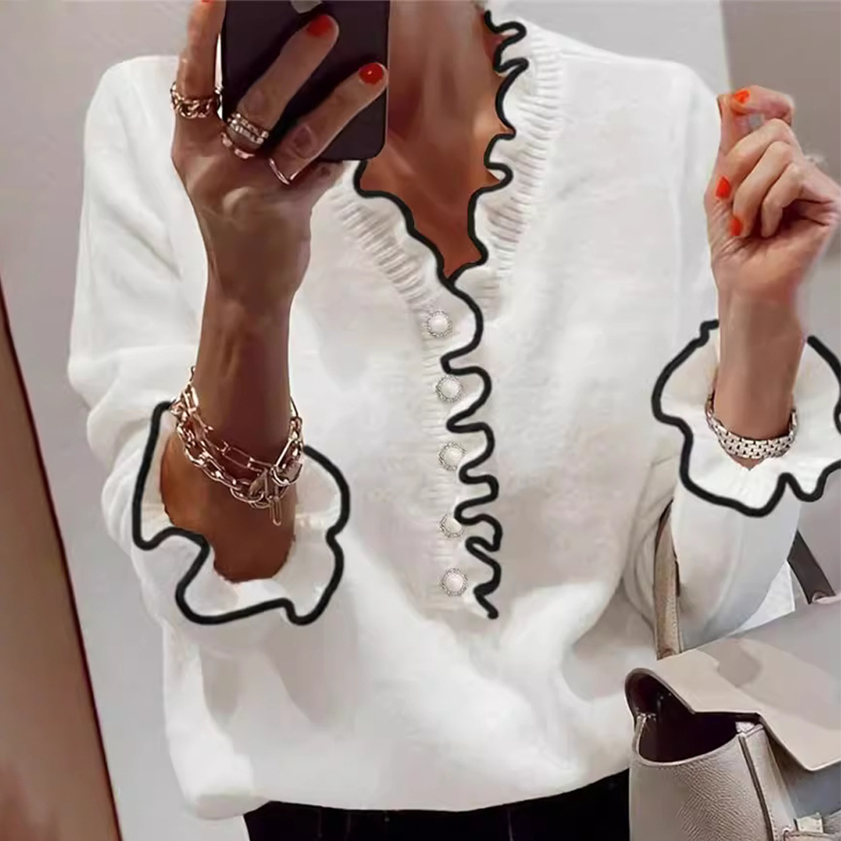 Femme en pull blanc à volants noirs, tenant un smartphone, portant des bracelets dorés, ongles rouges, sac à main beige. Mode élégante et moderne.