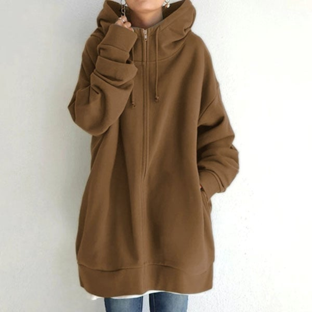 Sweat à capuche marron oversize, manches longues, porté par une personne debout contre un mur blanc, style décontracté et moderne.