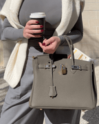 Femme tenant un sac à main en cuir gris avec cadenas doré, portant un café à emporter, vêtue d'un pull blanc et pantalon rayé, style chic urbain.