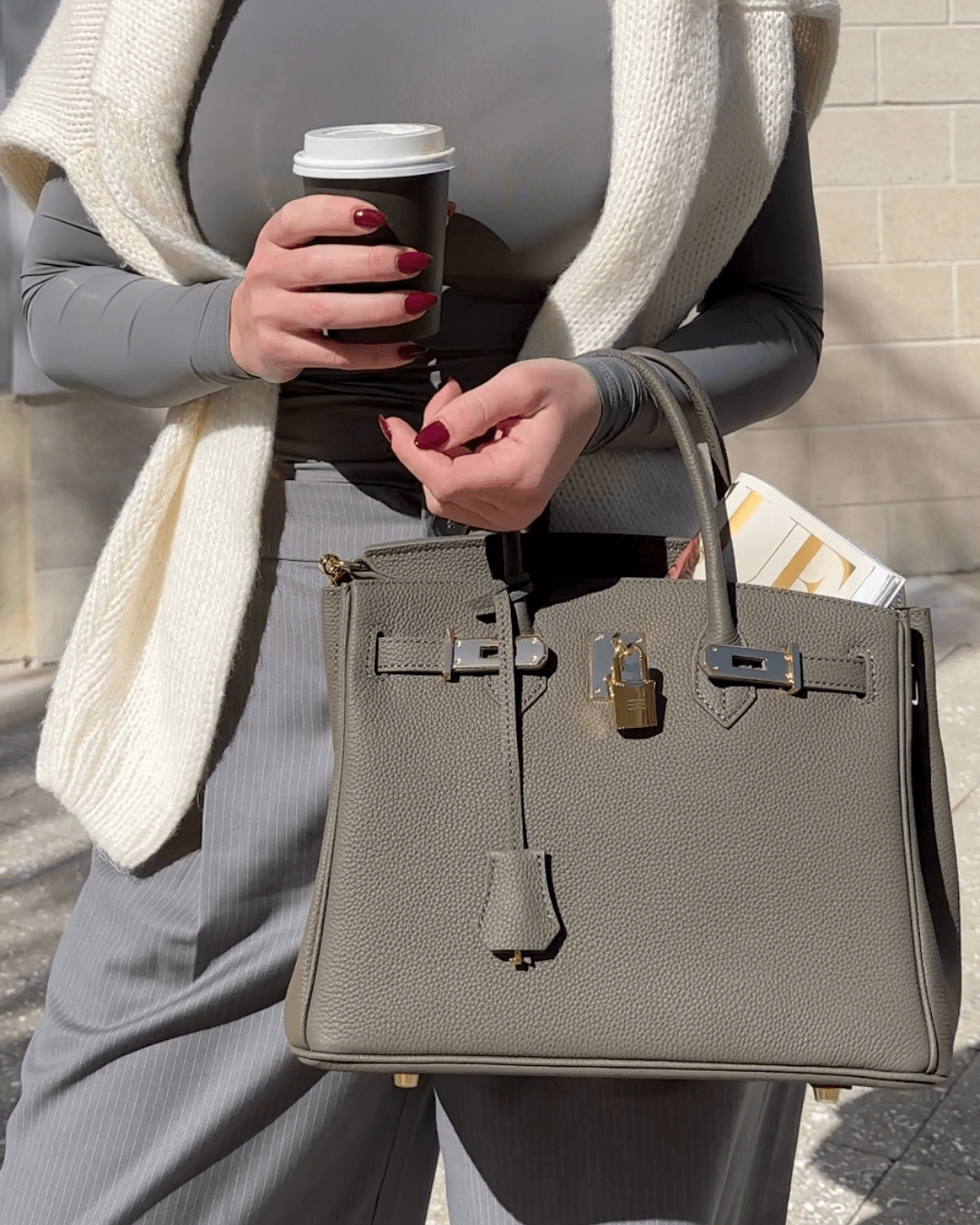 Femme tenant un sac à main en cuir gris avec cadenas doré, portant un café à emporter, vêtue d'un pull blanc et pantalon rayé, style chic urbain.