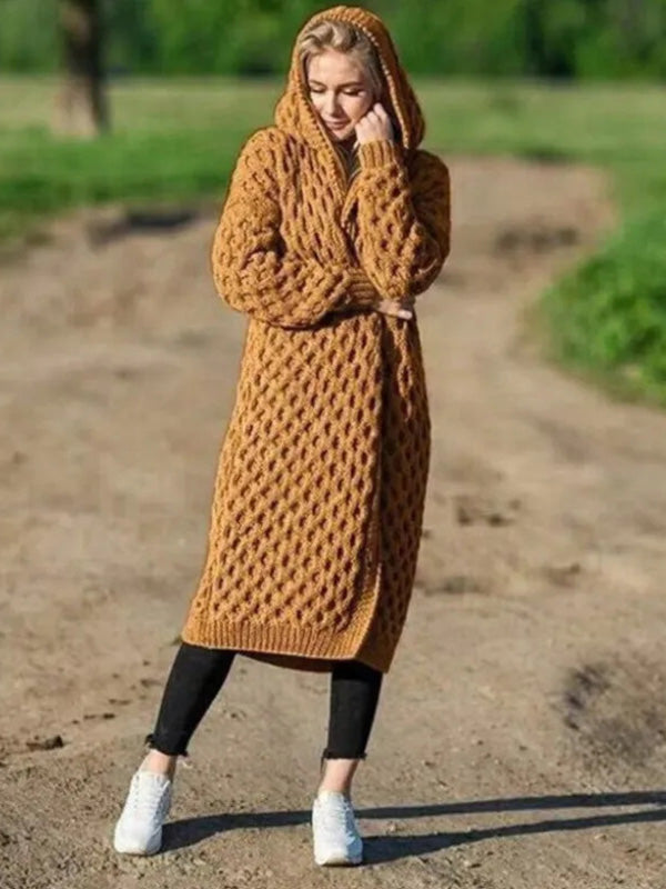 Femme portant un long cardigan en tricot marron à capuche, debout sur un chemin de terre, avec un fond de verdure. Mode automne, style décontracté.