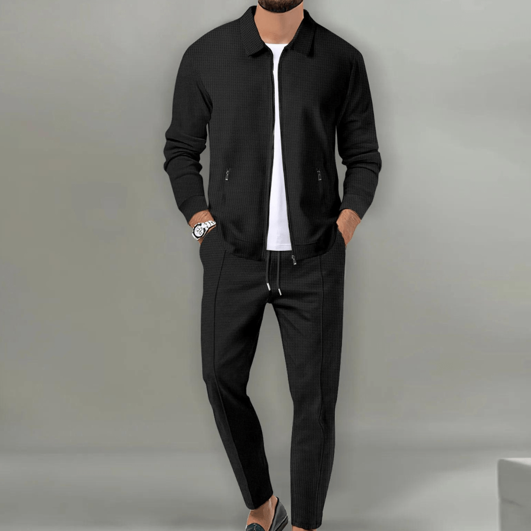 Homme portant un ensemble décontracté noir, veste et pantalon assortis, t-shirt blanc, montre argentée, chaussures grises. Mode masculine élégante.