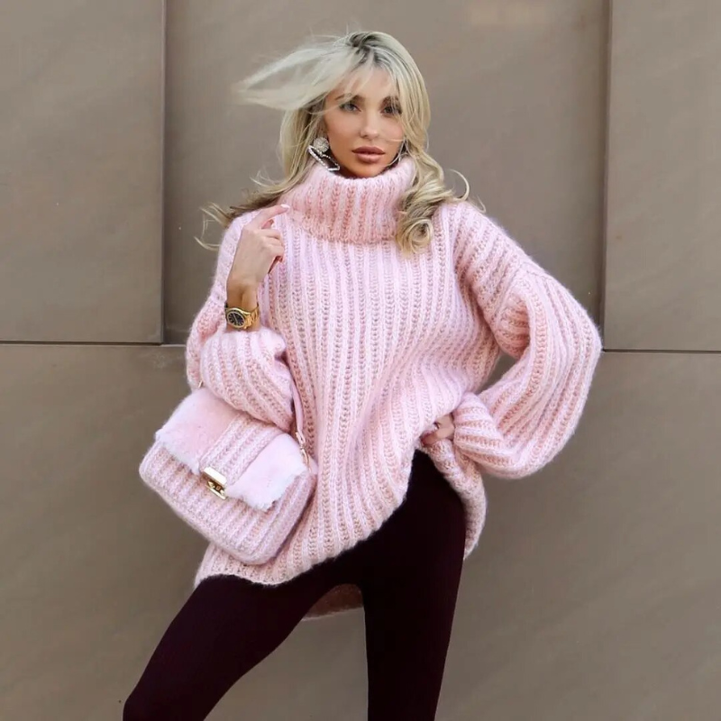 Femme en pull rose oversize, posant avec un sac assorti, devant un mur beige. Mode automne, style décontracté, tendance féminine élégante.