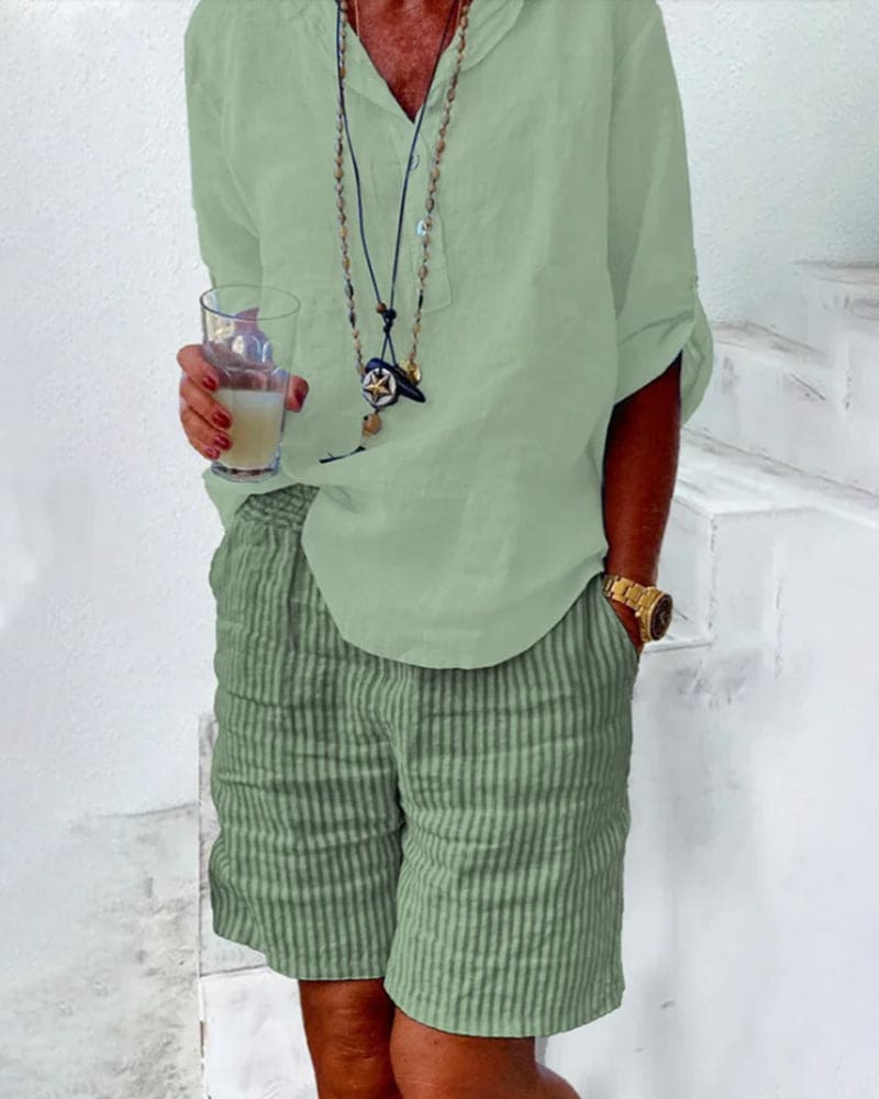 Homme en chemise verte et short rayé, tenant un verre. Style décontracté estival, accessoires tendance, montre dorée. Mode masculine élégante.