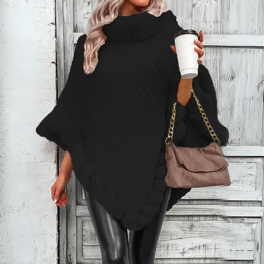Femme en poncho noir tricoté, tenant un gobelet de café et un sac à main beige, posant devant une porte en bois. Mode automne-hiver élégante.