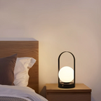 Lampe de chevet moderne avec abat-jour sphérique lumineux sur table en bois, à côté d'un lit avec oreillers, dans une chambre minimaliste.