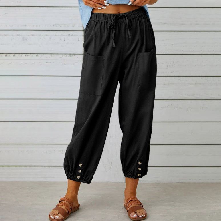Pantalon noir ample pour femme avec poches avant, boutons décoratifs aux chevilles, porté avec des sandales marron sur fond de planches en bois.