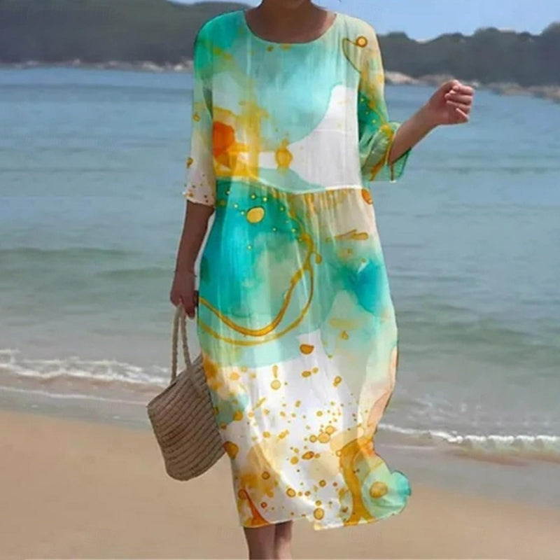 Robe longue d'été colorée avec motifs aquarelle, portée par une femme sur la plage, tenant un sac en osier. Mode plage, style décontracté.