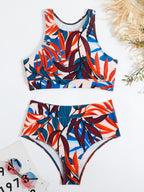 Maillot de bain deux pièces à motifs tropicaux colorés, avec feuilles rouges et bleues, lunettes de soleil et chapeau en paille en arrière-plan.