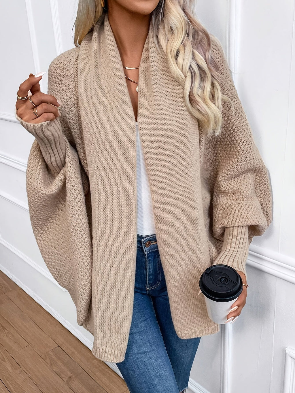 Femme portant un cardigan oversize beige, tenant un gobelet de café, posant contre un mur blanc. Mode automne, style décontracté, look chic.