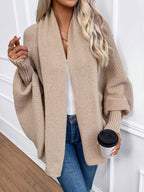 Femme portant un cardigan oversize beige, tenant un gobelet de café, posant contre un mur blanc. Mode automne, style décontracté, look chic.