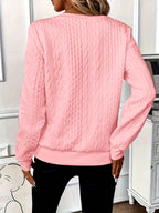Femme portant un pull rose en tricot torsadé, manches longues, dans un intérieur moderne. Mode féminine, tendance automne-hiver, confort élégant.