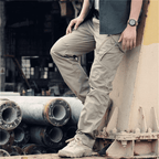 Homme portant un pantalon cargo beige, debout près de tuyaux industriels, montre-bracelet visible, style décontracté urbain, mode masculine moderne.