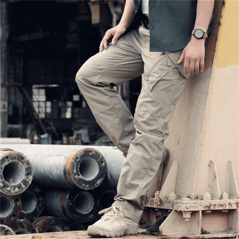 Homme portant un pantalon cargo beige, debout près de tuyaux industriels, montre-bracelet visible, style décontracté urbain, mode masculine moderne.