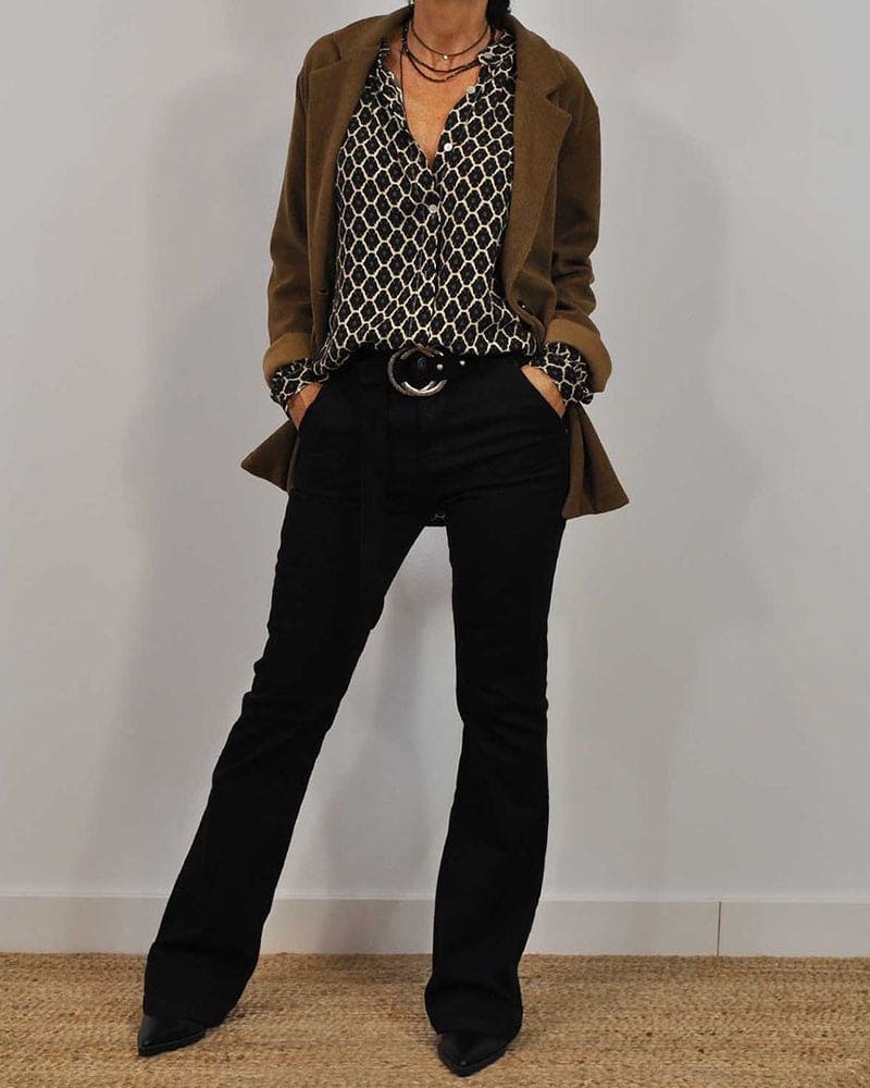 Femme portant un blazer marron, chemisier à motifs géométriques noir et blanc, pantalon noir évasé, ceinture noire, posant debout.
