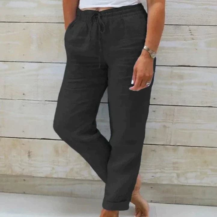 Pantalon noir décontracté pour femme, taille élastique avec cordon, poches latérales, porté devant un mur en bois clair. Mode confortable et élégante.
