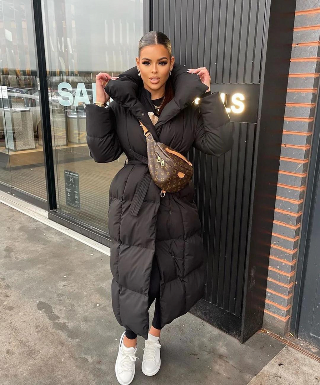 Jeune femme portant un manteau long noir, sac banane de luxe, posant devant une vitrine. Style hivernal chic, mode urbaine tendance.