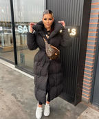 Jeune femme portant un manteau long noir, sac banane de luxe, posant devant une vitrine. Style hivernal chic, mode urbaine tendance.