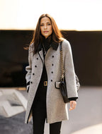 Femme élégante portant un manteau gris double boutonnage, sac à main noir à chaîne dorée, marchant en extérieur. Mode automne chic et tendance.