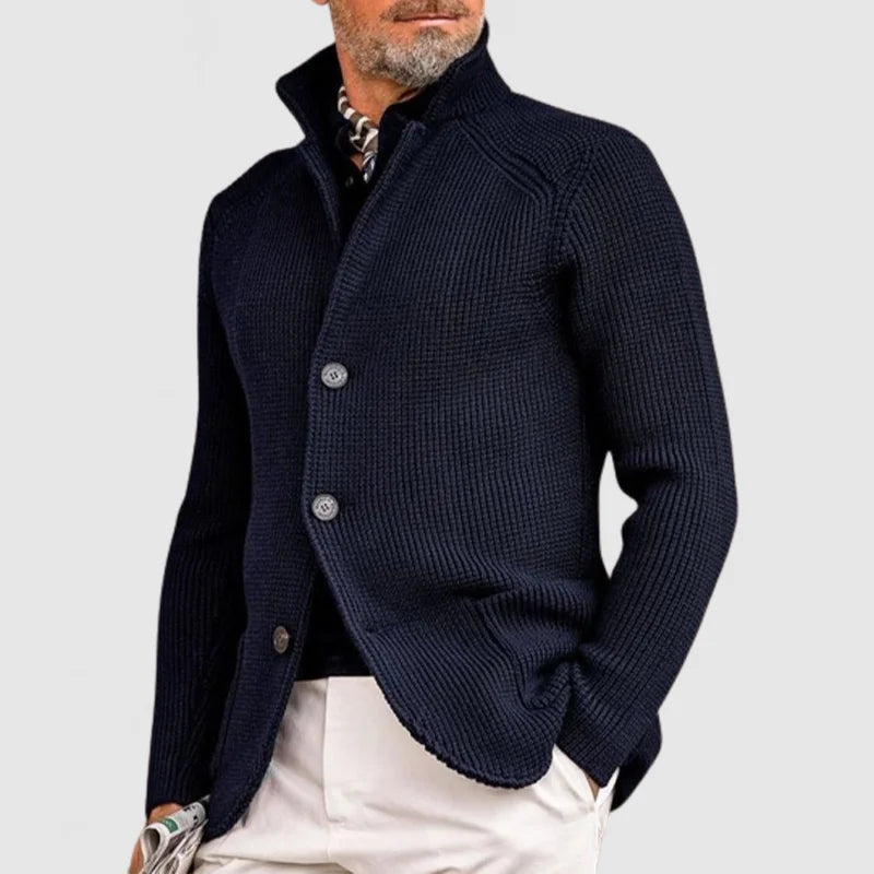 Homme portant un cardigan bleu marine élégant, col relevé, boutons visibles, sur chemise à motifs, pantalon beige. Mode masculine chic et moderne.