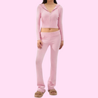 Femme en ensemble de survêtement rose, comprenant un sweat à capuche zippé et un pantalon taille basse, posant sur fond rose. Mode décontractée féminine.