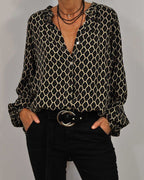 Chemisier à motifs géométriques noir et beige, manches longues, col ouvert, porté avec un pantalon noir et ceinture boucle argentée.