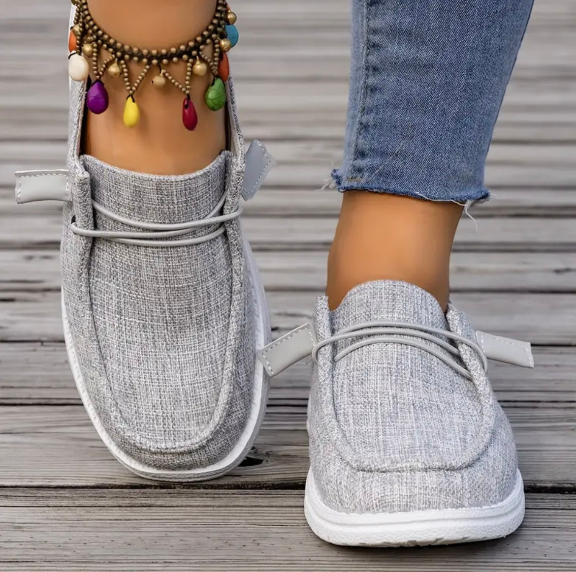 Chaussures décontractées en toile grise pour femme, portées avec un jean ajusté et un bracelet de cheville coloré, sur fond de plancher en bois.