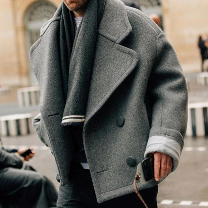 Homme portant un manteau gris oversize et une écharpe, tenant un smartphone. Mode automne-hiver, style urbain élégant.