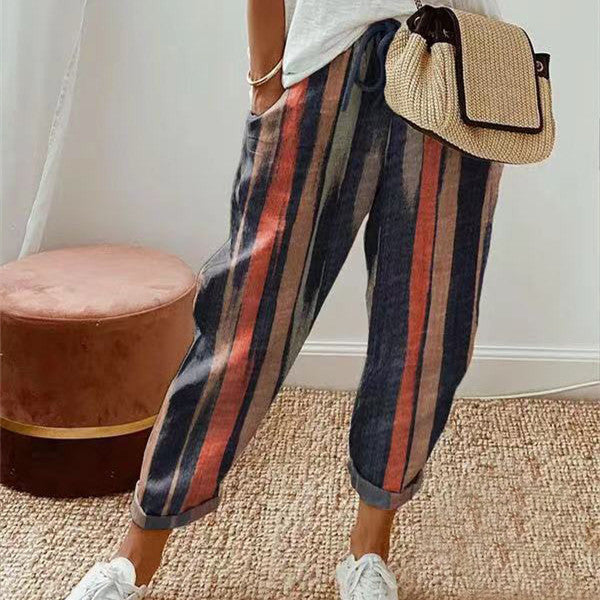 Pantalon à rayures multicolores pour femme, style décontracté, avec poches. Tapis en jute et pouf rose en arrière-plan. Mode tendance.