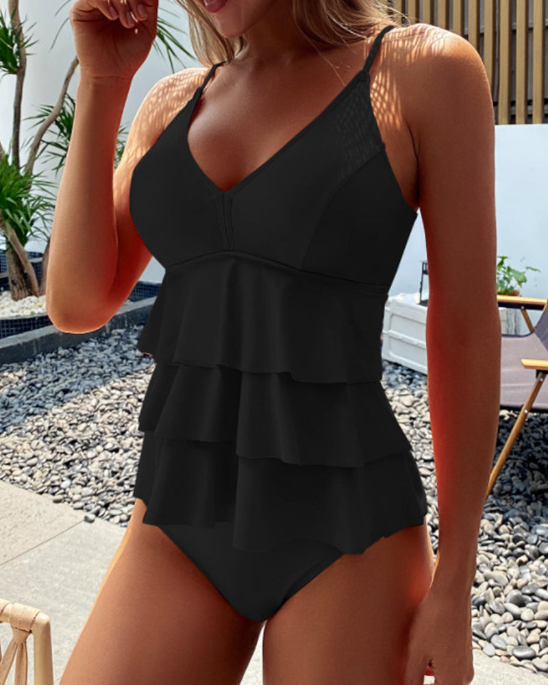 Femme en maillot de bain noir à volants, posant près de plantes et mobilier extérieur. Mode plage élégante, tenue d'été tendance.