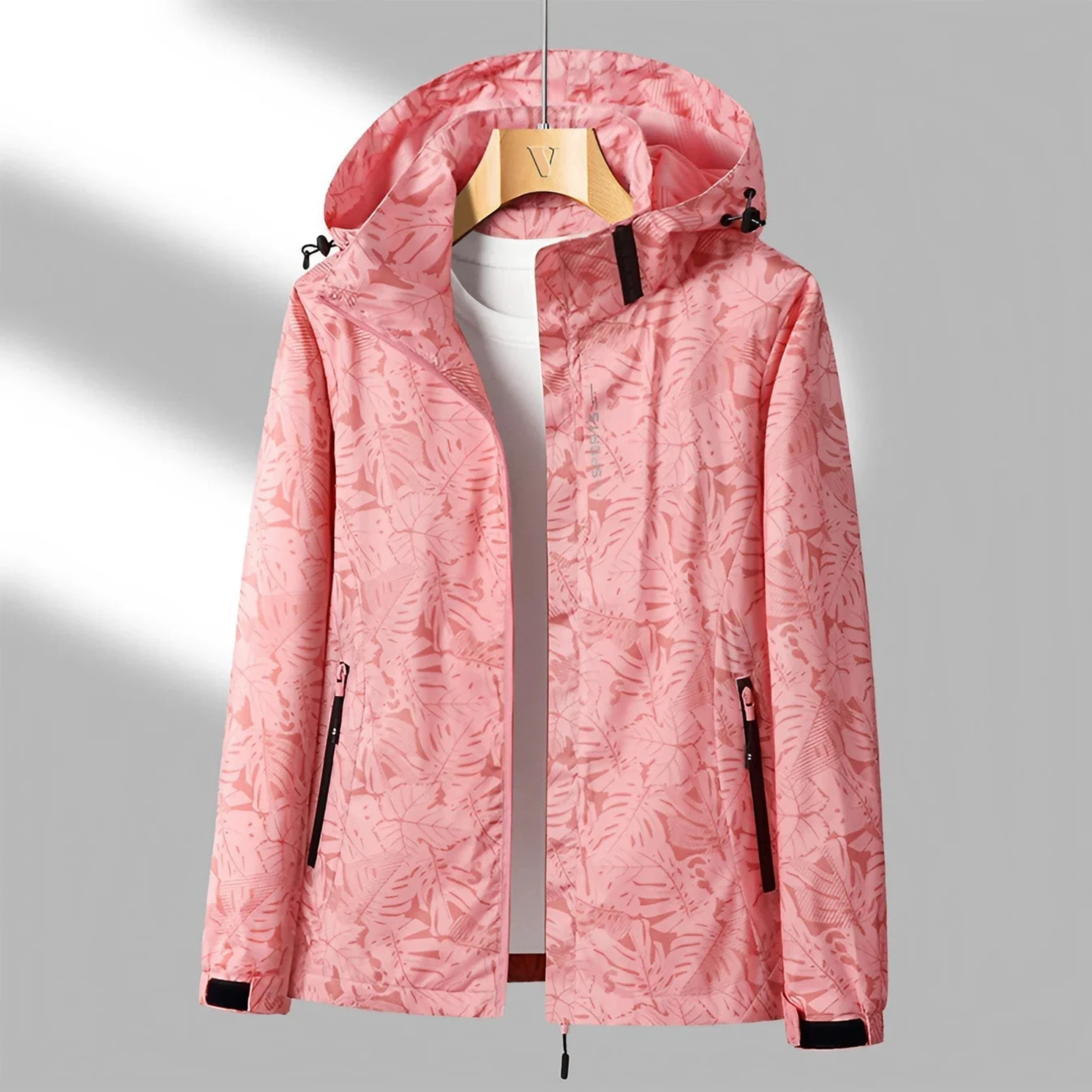Veste imperméable rose à motifs floraux, capuche ajustable, fermetures éclair noires, suspendue sur un cintre en bois, fond gris clair.