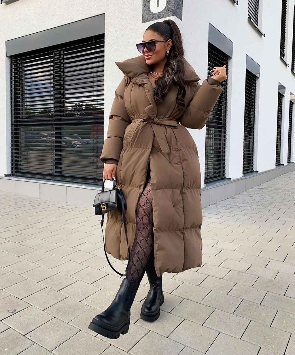 Femme portant un manteau long marron, collants à motifs, bottes noires et lunettes de soleil, tenant un sac à main noir devant un bâtiment moderne.