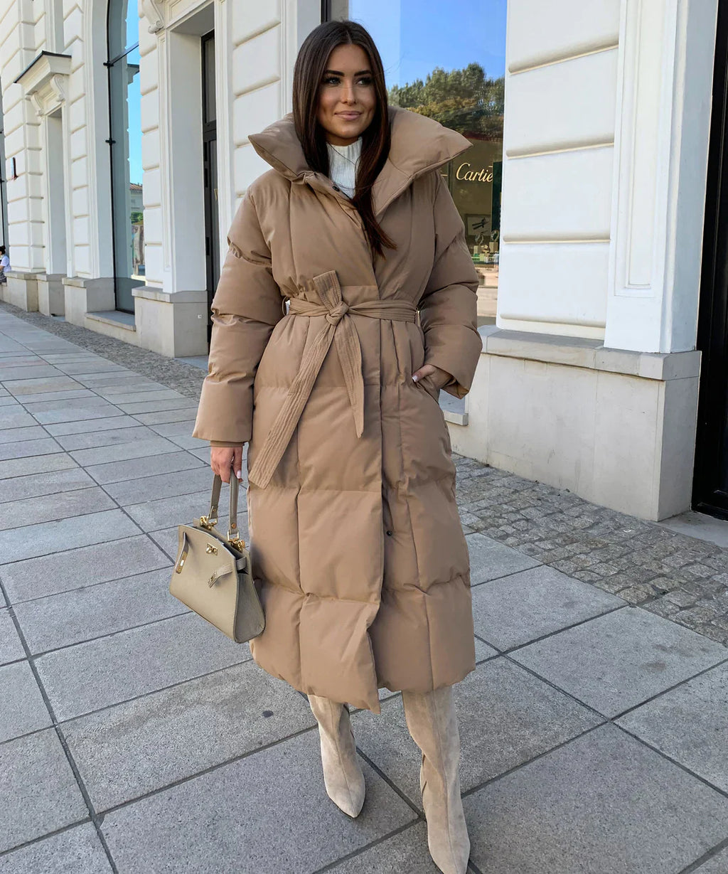 Femme portant un manteau long matelassé beige, bottes hautes grises et sac à main élégant, marchant sur un trottoir urbain devant des boutiques.