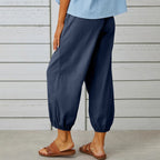 Pantalon ample bleu marine pour femme, style décontracté, avec ourlets élastiques, porté avec des sandales marron, devant un mur en bois blanc.