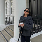 Femme élégante en manteau gris, lunettes de soleil noires, tenant un café devant un bâtiment classique blanc avec colonnes. Mode urbaine chic.