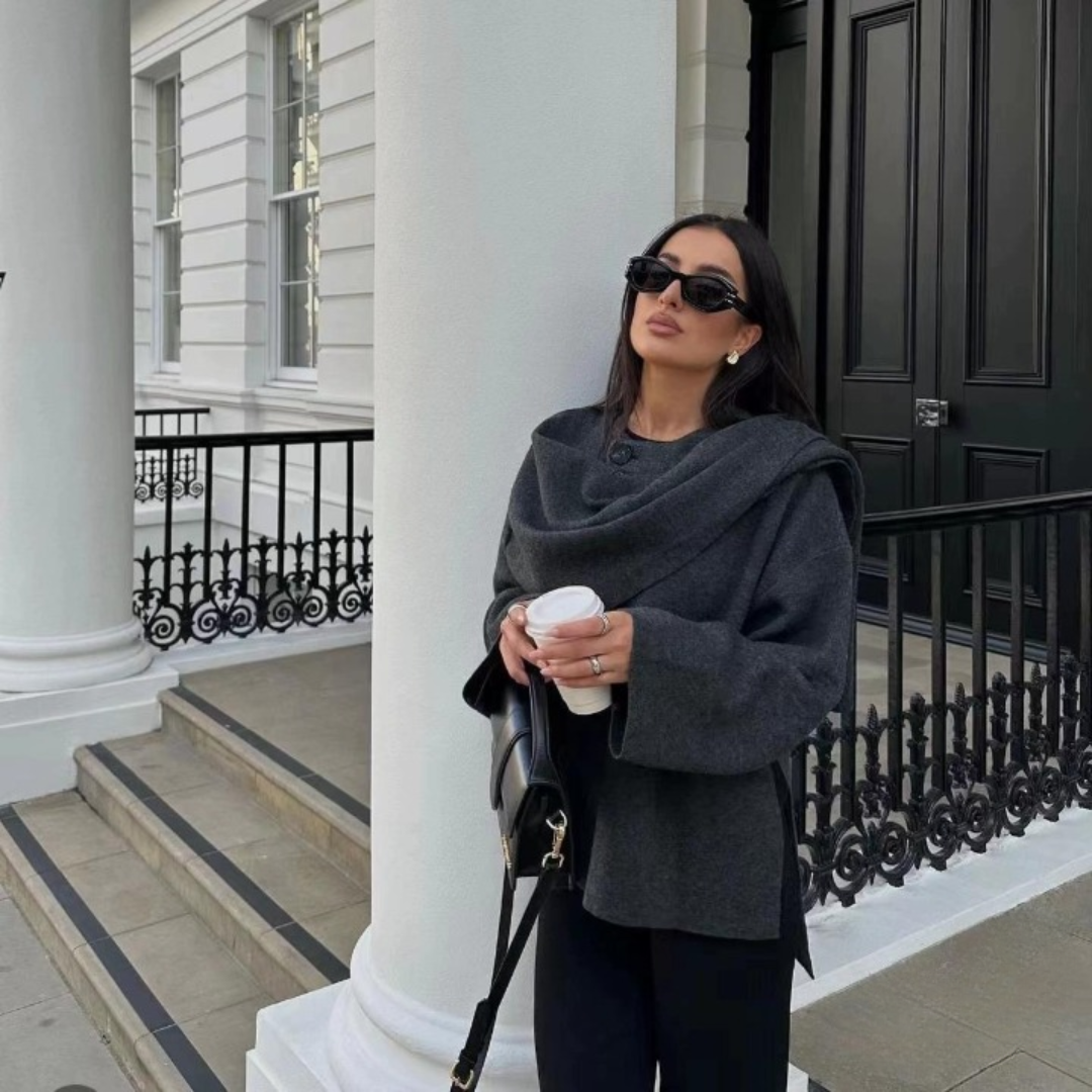 Femme élégante en manteau gris, lunettes de soleil noires, tenant un café devant un bâtiment classique blanc avec colonnes. Mode urbaine chic.