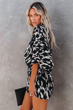 Jeune femme blonde portant une robe à motifs noir et blanc, tenant une pochette noire, posant devant un mur gris. Mode féminine élégante.