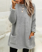 Femme portant un pull en laine gris avec poches, col rond, manches longues, collier doré, posant dans un intérieur lumineux. Mode hiver tendance.