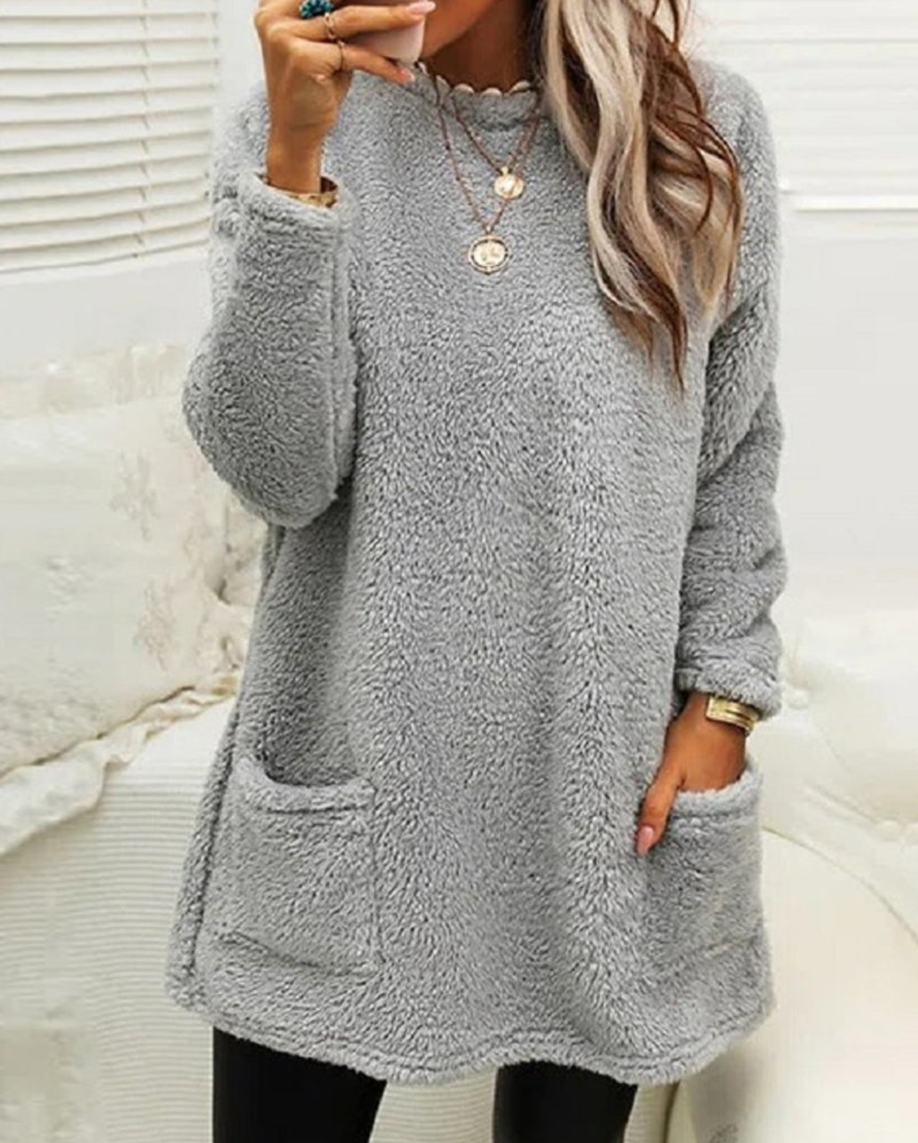 Femme portant un pull en laine gris avec poches, col rond, manches longues, collier doré, posant dans un intérieur lumineux. Mode hiver tendance.