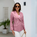 Femme élégante portant un blazer rose à boutons dorés, lunettes de soleil, posant dans un intérieur moderne avec plante verte. Mode chic et tendance.