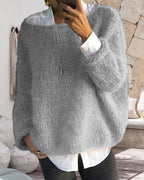 Personne portant un pull en laine gris ample, sur une chemise blanche, dans un intérieur moderne avec murs en pierre et décoration minimaliste.