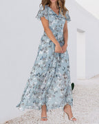 Robe longue fleurie bleue pour femme, manches courtes, col en V, portée avec des sandales argentées, fond blanc minimaliste.