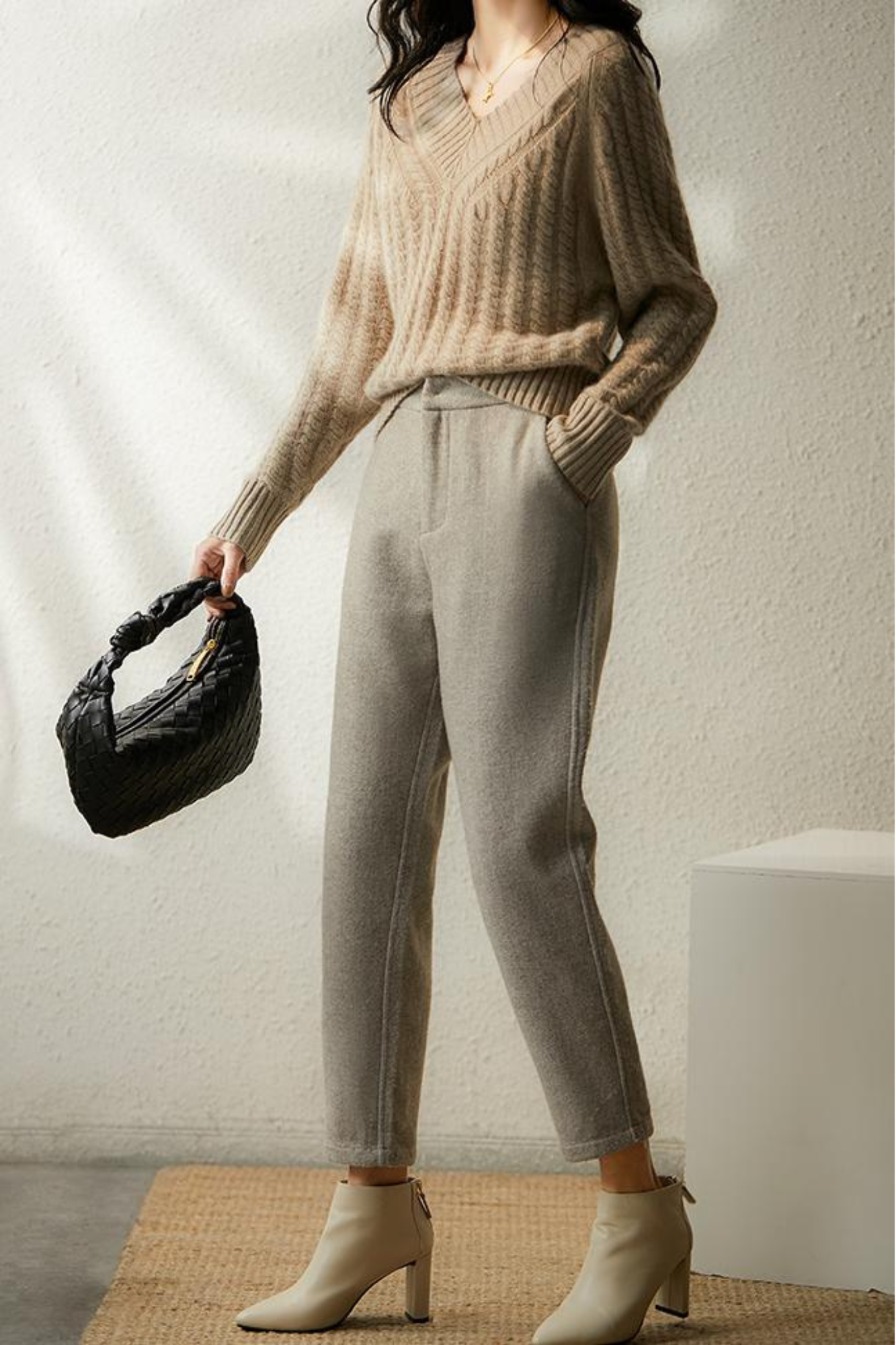 Femme en pull beige et pantalon gris, tenant un sac noir. Mode automne-hiver, style élégant et décontracté, chaussures à talons beiges.