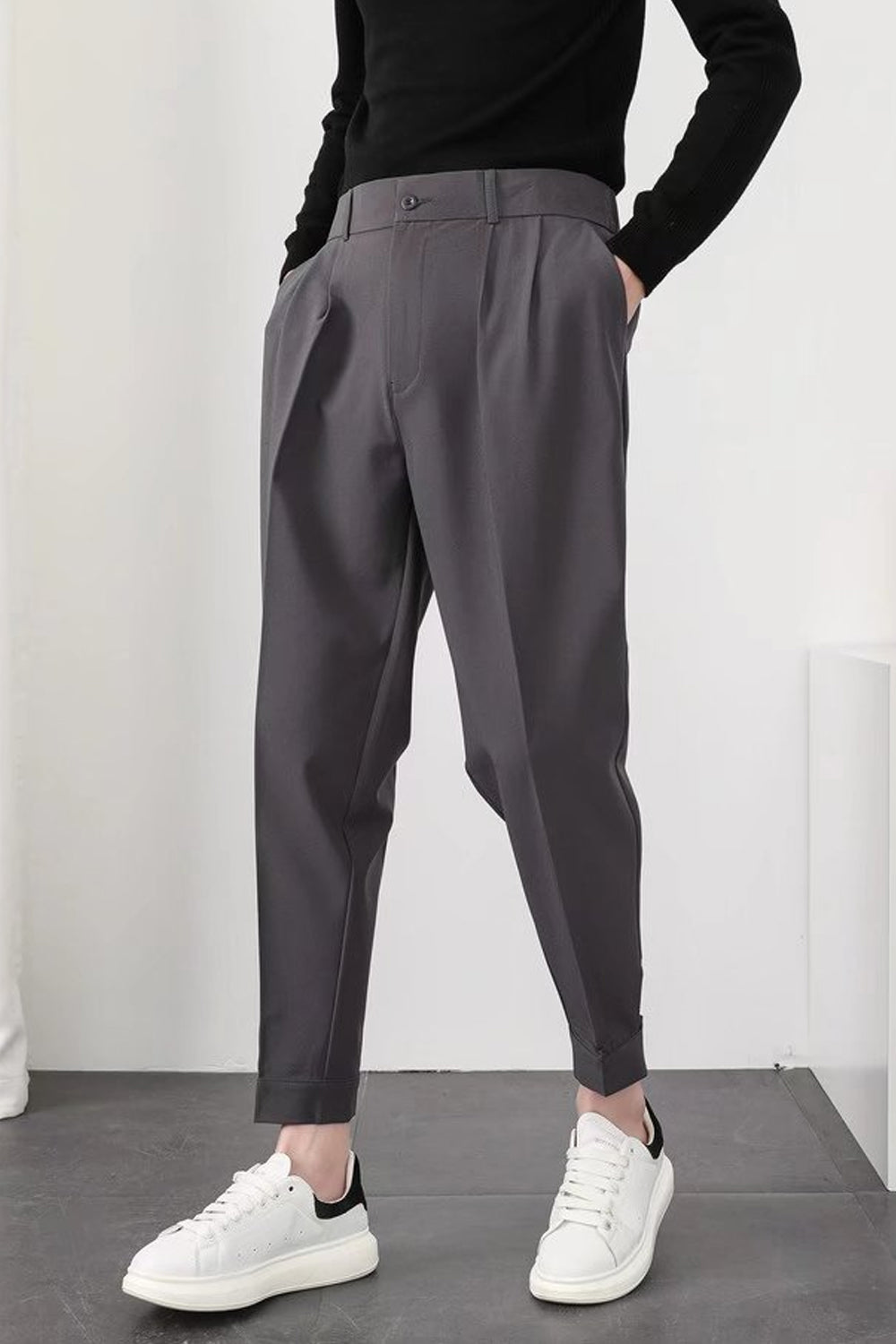 Pantalon gris homme élégant, coupe ample, avec pinces, porté avec pull noir et baskets blanches modernes. Mode masculine chic et décontractée.