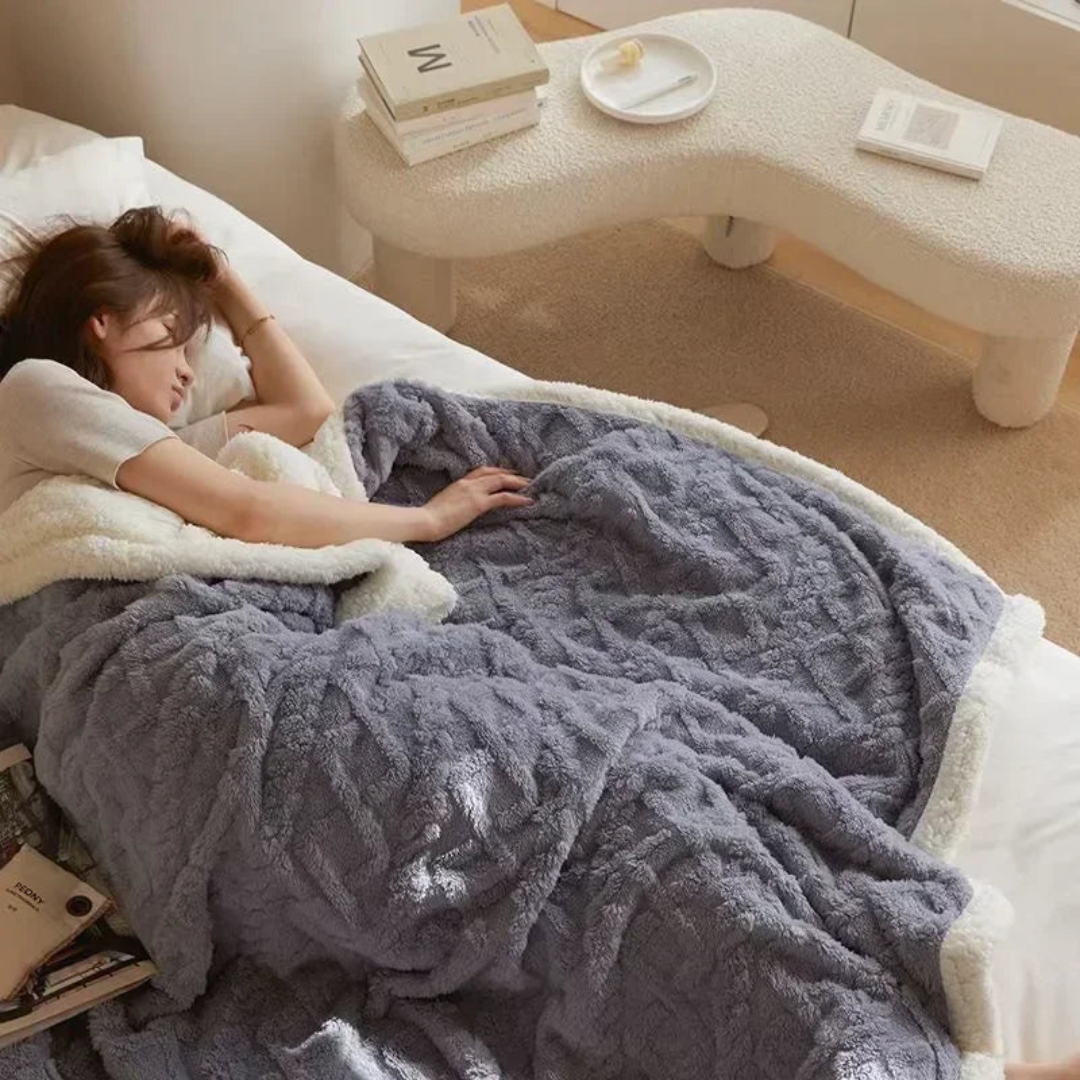 Femme dormant sous une couverture en fausse fourrure grise sur un lit, avec des livres à côté. Chambre lumineuse et moderne. Confort et détente.