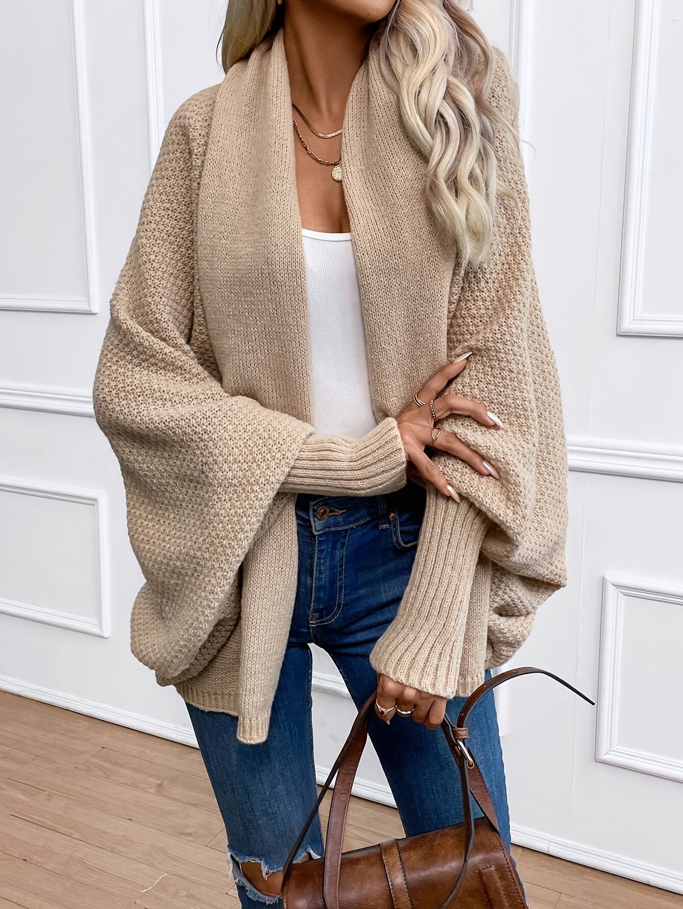 Femme portant un cardigan beige ample, style décontracté, avec un top blanc et un jean bleu, tenant un sac marron. Mode automne-hiver tendance.