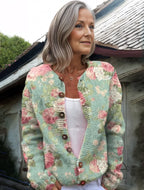 Femme portant un cardigan en laine à motifs floraux roses et verts, devant une maison rustique. Mode automne, style vintage, élégance décontractée.