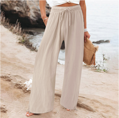 Pantalon large beige femme sur plage, style décontracté, avec sac en paille. Mode estivale, tenue confortable, bord de mer.