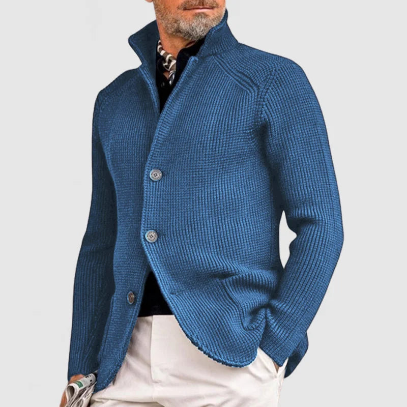 Homme portant un cardigan bleu élégant, col relevé, boutons visibles, sur chemise noire et pantalon beige. Mode masculine chic et moderne.